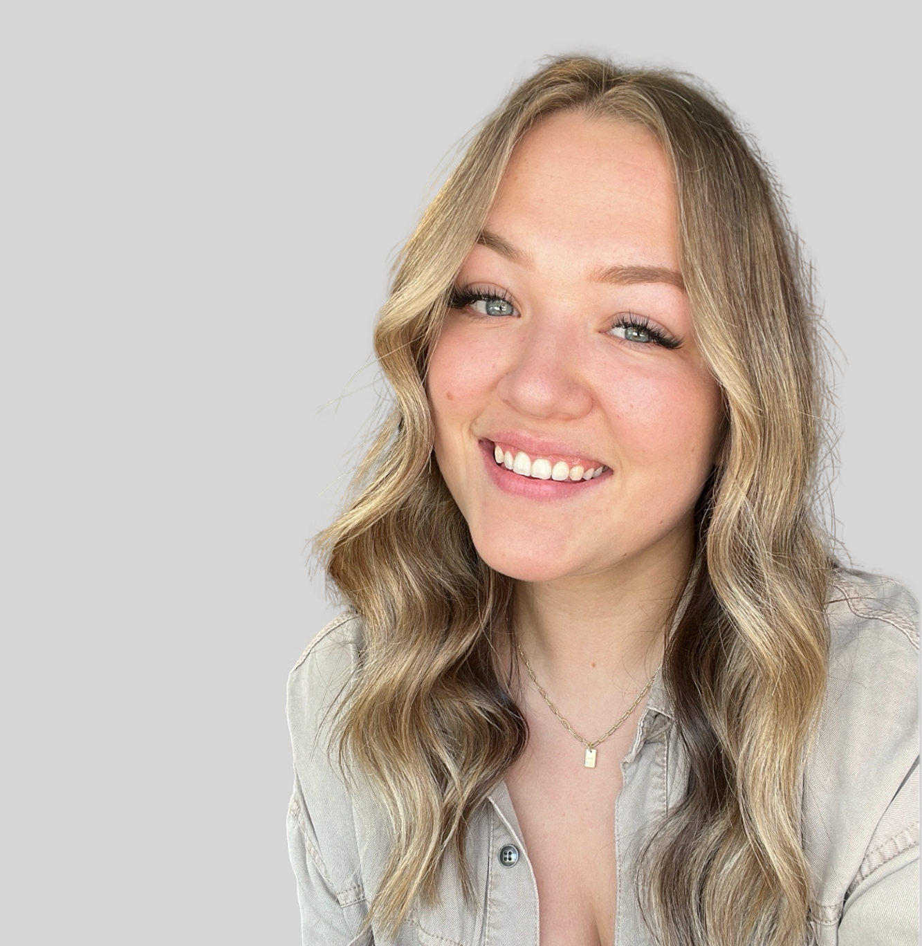 Smiling young woman with wavy blonde hair, wearing a gray button-down shirt and a gold necklace, against a light gray background.