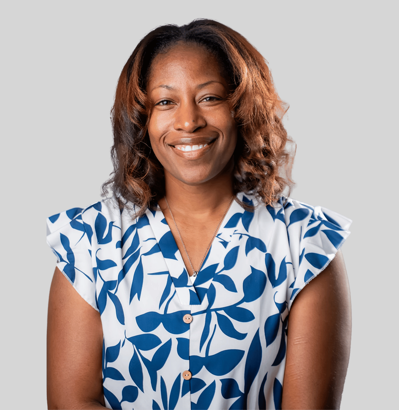 Smiling woman with shoulder-length wavy hair wearing a blue and white leaf-patterned blouse against a gray background.