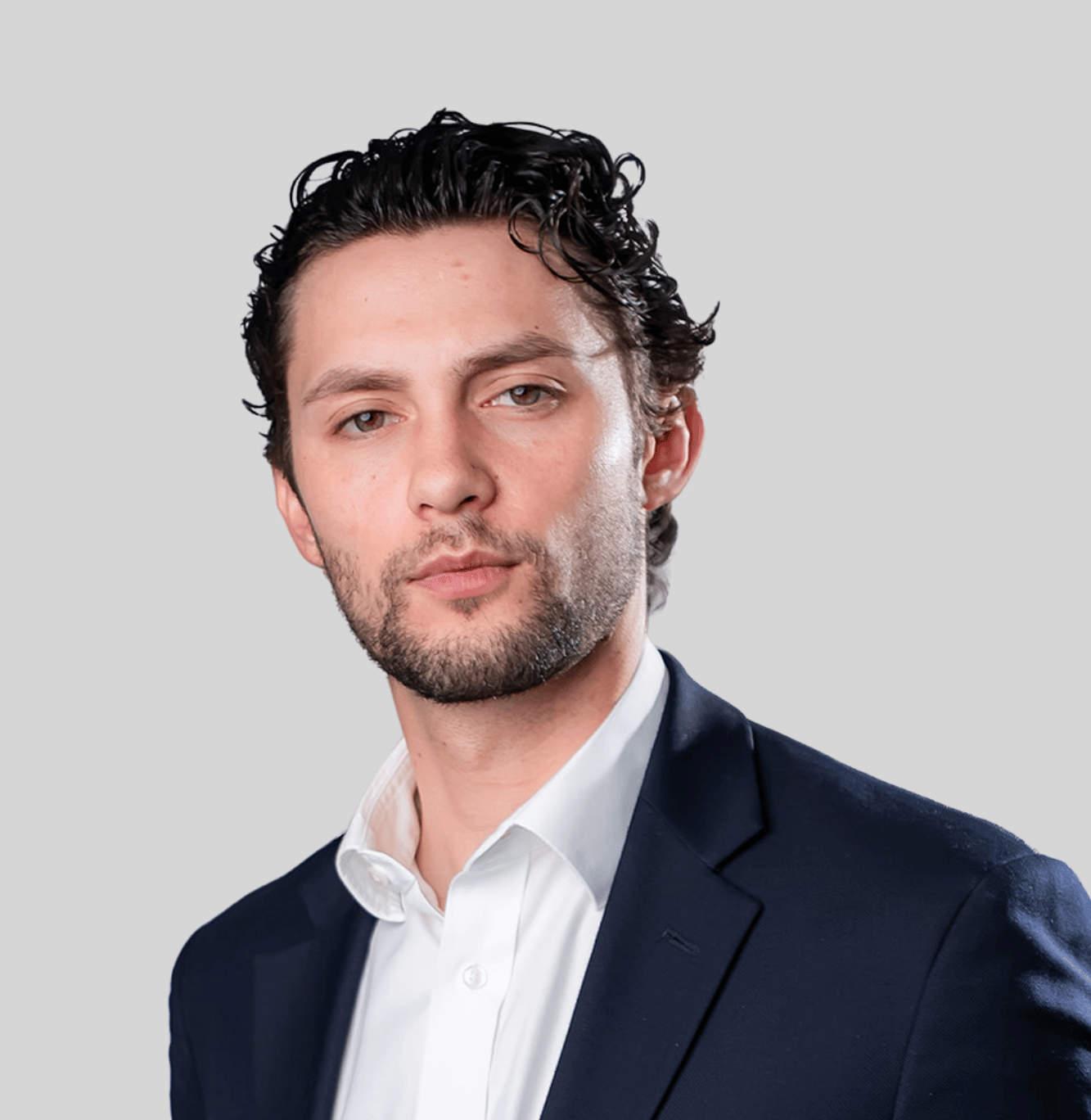 Portrait of a man in a navy blazer and white shirt, looking at the camera against a gray backdrop, presenting a professional headshot.