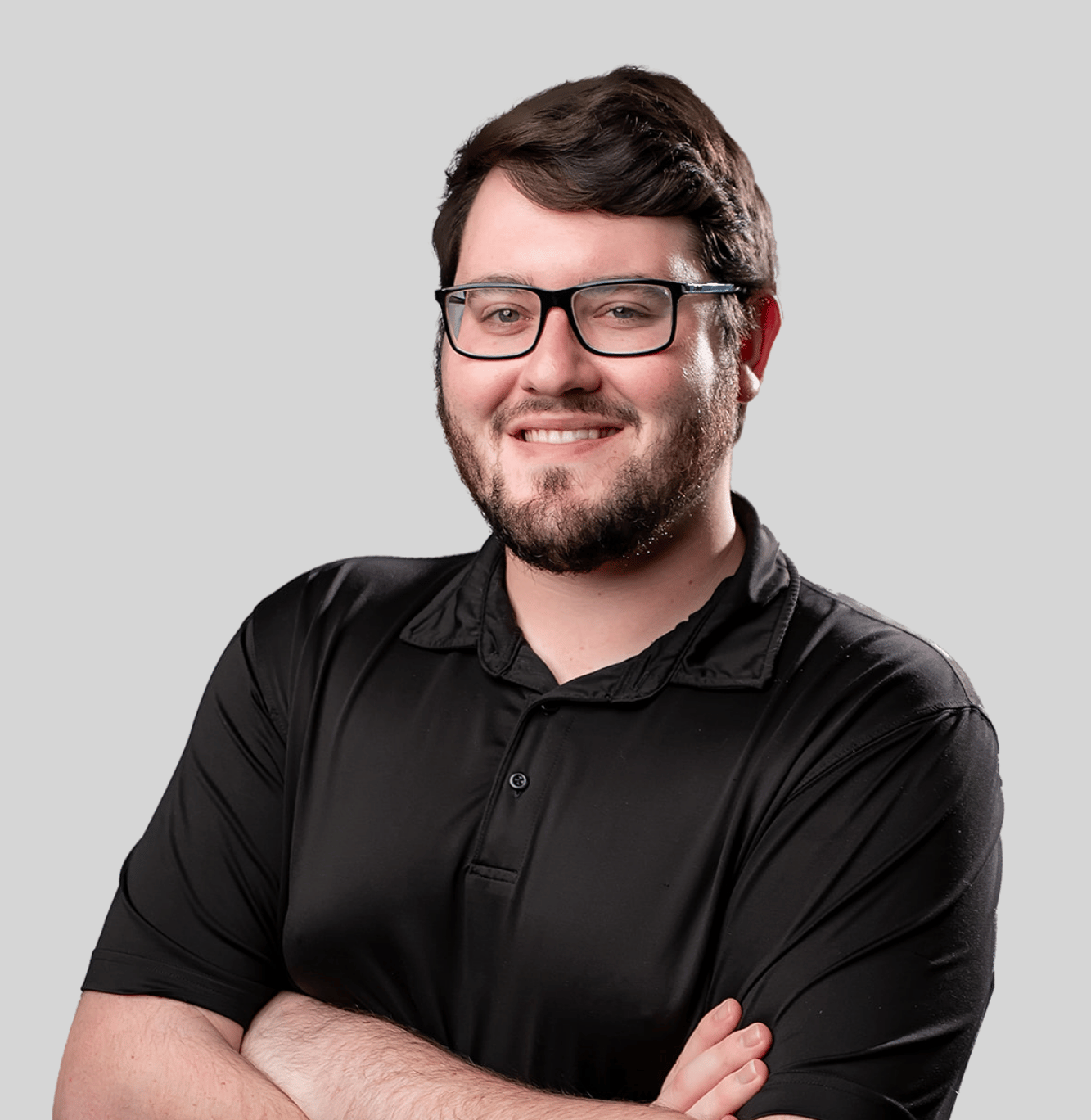 Smiling man with dark hair and glasses wearing a black polo, arms crossed against a light gray background.